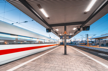 High speed train in motion on the railway station with beautiful illumination at night. Moving blurred modern intercity train on the railway platform at dusk. Passenger transportation. Railroad