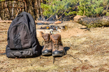 Backpack and touristic boots on a ground in a coniferous forest. Bonfire on a background