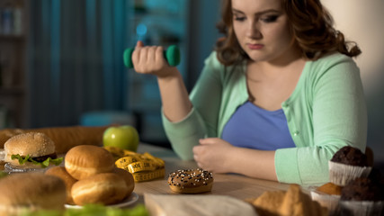 Overweight lady exercising and sadly looking at junk food, obesity problem