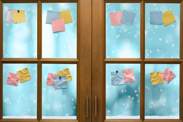 Wooden window frame with colorful blank sticker notes on glass and falling snow in freezing blue tone bokeh in background.