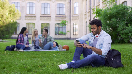 Hispanic male student sitting on grass and using cellphone, app for acquaintance