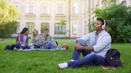 Naklejka na ściany i meble Handsome hispanic student sitting on grass and enjoying music in headphones