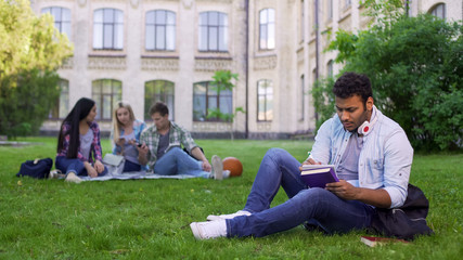Fototapeta na wymiar Serious biracial male student sitting on grass and writing essay, doing homework