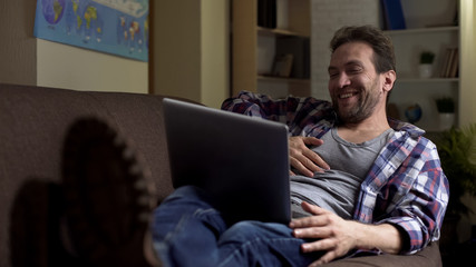 Lazy grubby male lying on couch, looking at laptop laughing at show on screen