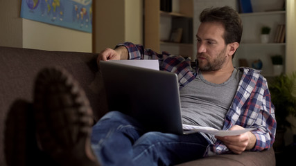 Male freelancer on couch looking at documents and working on laptop at home