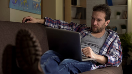 Tired adult male on couch looking at laptop concentrated, continuous job search