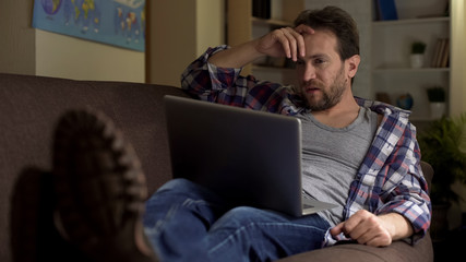 Guy sitting on sofa, looking at laptop, angry with received news, bitterness