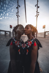 Vertical wide-angle shot of a charming black girl with braided hair, in dress, touching a glass wall with her face while standing outdoors; African girl with braids leaning against glass wall