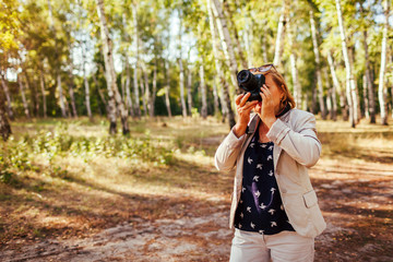 Middle-aged woman taking pictures using camera in autumn forest. Senior woman walking and enjoying hobby