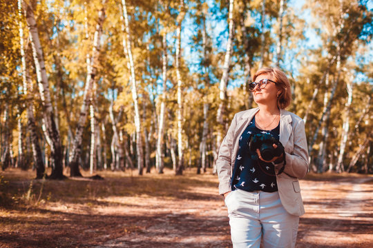 Middle-aged woman taking pictures using camera in autumn forest. Senior woman walking and enjoying hobby