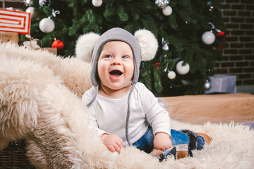 Fototapeta premium Subject children christmas new year. Caucasian little funny baby boy 1 year old sitting sleigh bear skin Christmas tree head warm hat with balabons balls, balls and jeans around box gifts in evening.