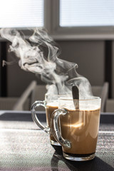 Two glass cups of coffee with milk are on the table. Steam close up