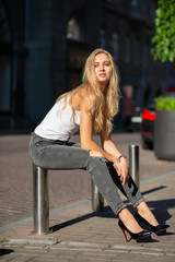 Seductive blonde model with long hair wearing jeans and t shirt posing at the city in sun rays