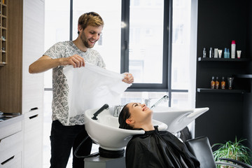 Charming young woman smiling happily while professional male hairstylist preparing her hair for the haircut drying it with a towel
