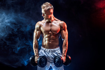 Fototapeta na wymiar Topless man exercising biceps with dumbbells posing in studio full of colored smoke