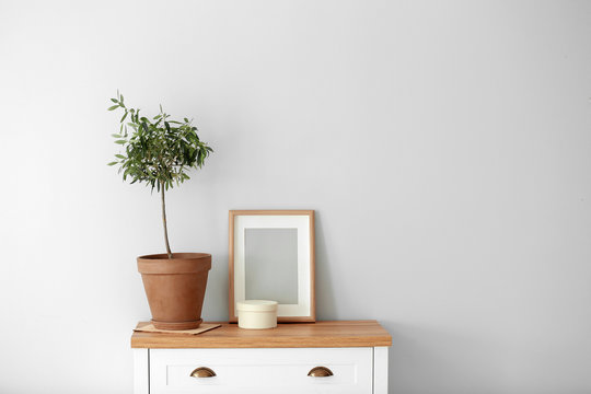 Olive tree in brown pot on chest of drawers near wall. Space for text