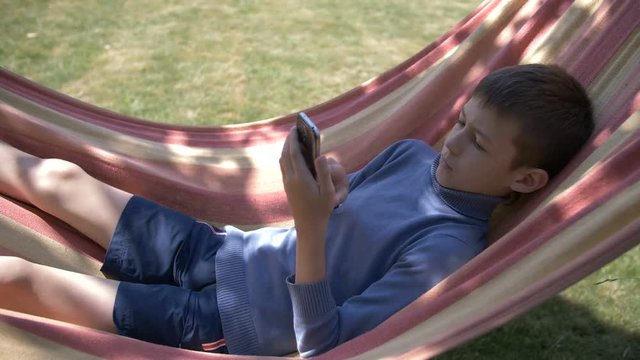 boy lies on a hammock, uses a phone