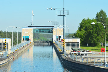 View across the top of a filled and closed lock. Some ducks are swimming on the water. A control tower arches over the lock gate, which is closed. The sky is blue.