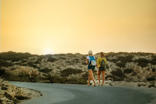 Two young tourists with backpacks walking on road and enjoying sunset. Concept travel trekking, hiking in mountains or sea in Cyprus.