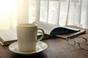 Coffee cup on wooden table by the window with opening book and eye glasses as background. Soft morning light shines through the curtain.