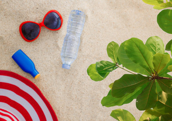 Sunscreen, hat, sunglasses and bottle of water on sand beach under green leaves in soft and blur background