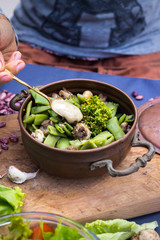 Woman hand adds sesame seeds tahini dip sauce with spoon to green beans, broccoli, mushrooms with spices. Cooked vegetables in copper vintage pot. Vegan vegetarian healthy food.