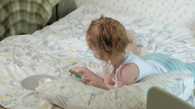 A little beautiful girl cheerfully makes a cucumber masks at home and uses a smartphone