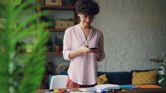 Smiling curly-haired lady is taking pictures of desk creating flat lay using smartphone camera standing in modern apartment. Modern technology and design concept.