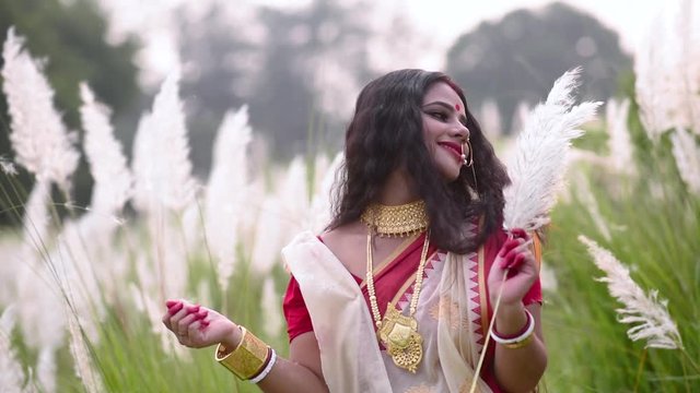 A married happy and elegant Indian woman is playing with kaash phool white flowered grass on a windy day, sunset or sunrise. slow motion