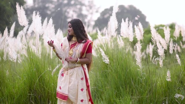 A calm and beautiful Indian woman is happy and plays with white grass called saccharum spontaneum in a field, gold jewellery, slow motion