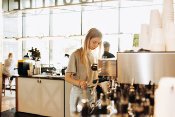 A young beautiful thin blonde,dressed in casual outfit,is cooking coffee in a popular coffee shop.