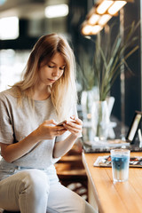 A cute thin blonde girl,dressed in casual style,drinks coffee and looks at her phone in a coffee shop.