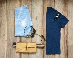 Fashionable concept, men’s clothes with sling bag leather, blue shirt , sunglasses, denim shorts and earphone on wooden background. pack shots, top view