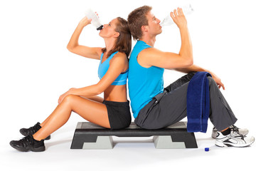 Active sports couple sitting back to back on aerobic stepper drinking water from bottle after exercise session over white background