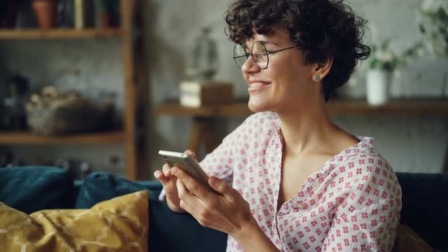 Happy girl is using smartphone looking at screen and laughing texting friends and checking social media accounts in internet sitting on sofa at home.