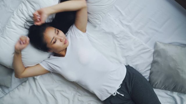 Pretty Asian lady is sleeping on bed then opening eyes and stretching her arms with smile. Soft pillow and white linen blanket and sheets are visible.