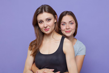 Two attractive young women standing embracing, one besides another one. Dark hair, blue and brown eyes. Isolated, studio, models, copy space.