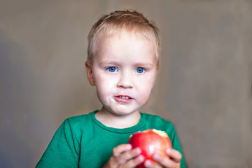 Little cute caucasian boy with blue eyes and blonde hair in green t-short eats red apple by himself. The apple is on his hands, slightly blurred. Indoors, close up