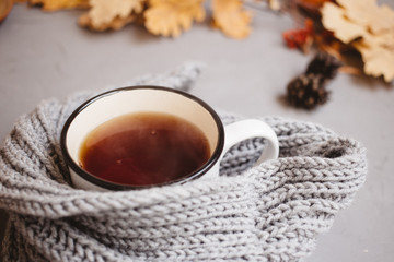 orange and yellow autumn leaves, a Cup of tea wrapped in a warm knitted scarf and Notepad with handle on gray background, top view, with copy space
