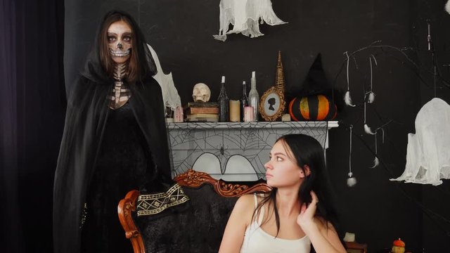 Attractive afraid brunette scared by creepy woman in Death Reaper cape Halloween costume in beautiful decorated room