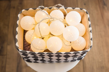 Christmas lights garland with round white lanterns in a gray box on a white stand on a wooden floor. Preparing for Christmas and New Years concept. Apartment decoration. Top view