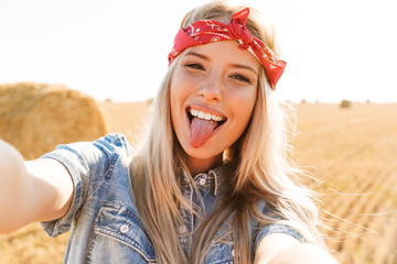 Portrait of amusing girl 20s laughing and taking selfie while walking through golden field, during sunny day
