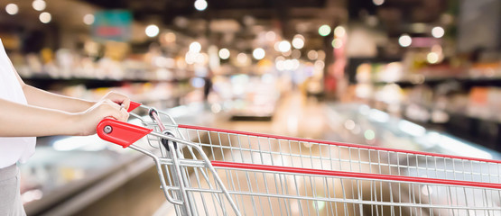 Woman hand hold empty red shopping cart with abstract blur supermarket discount store aisle...