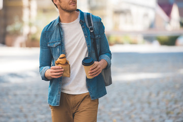 cropped shot of happy man with backpack, coffee to go and croissant walking by street and looking away