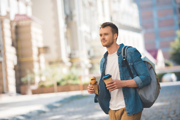 handsome young man with backpack, coffee to go and croissant walking by street and looking at camera