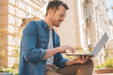 happy young man with paper cup of coffee using laptop on street