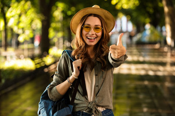 Happy woman walking outdoors with backpack showing thumbs up.