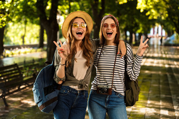 Beautiful young happy women friends walking outdoors with backpacks showing peace gesture.