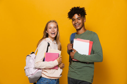 Photo of happy students guy and girl 16-18 wearing backpacks smiling and holding exercise books, isolated over yellow background