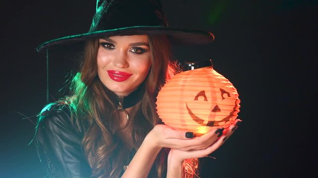 Halloween. Sexy witch girl holding pumpkin lantern. Beautiful young woman in witches hat with long curly red hair and bright makeup. 4K UHD video 3840X2160